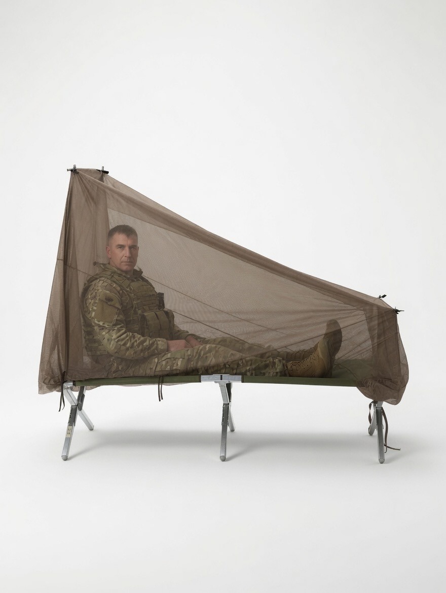 A soldier in camouflage clothing relaxes inside a mesh mosquito net, set up over a folding cot, highlighting camping or field readiness.